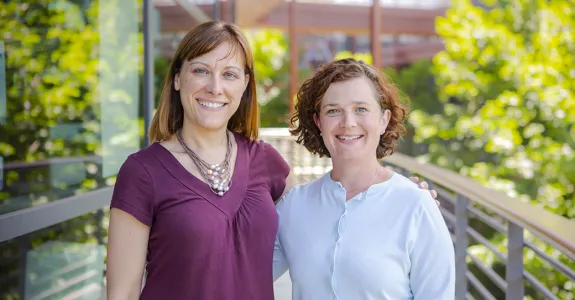 Outdoor photo of two female faculty members standing on a walkway with their arms around each other, smiling.