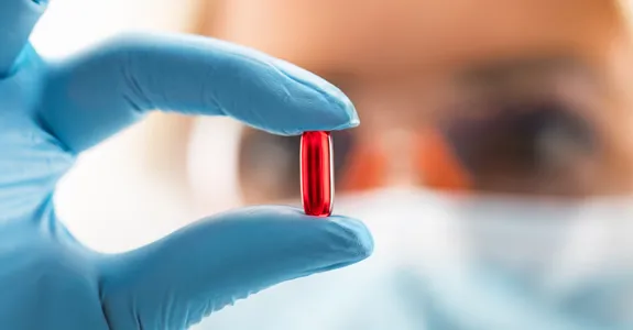 Photo of young female researcher wearing blue gloves and safety glasses, holding up a translucent red pill.