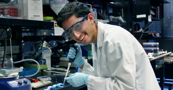 Photo of high school-aged researcher Rohan Mehrhotra in the wet lab space using a micropipette and wearing a lab coat and safety goggles.