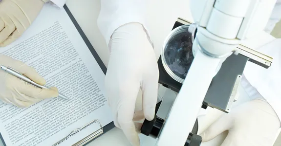 Photo from above of two scientists wearing gloves: one works with a microscope and one has a typed report on a clipboard.