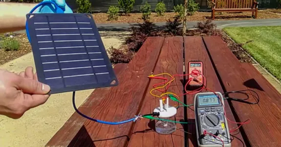 Photo of a solar panel being held up by a researcher's hands, with panel connected to a glass container and multiple sensors displaying readouts, via long wires.