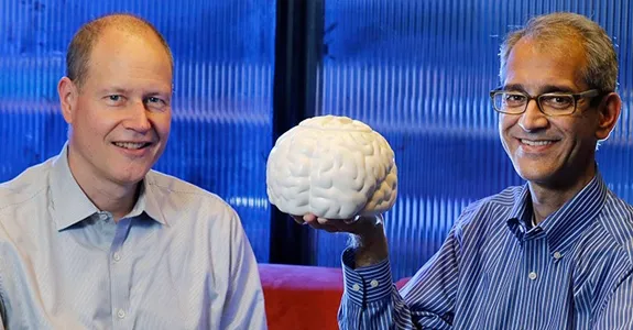 Photo of Dr. Jaime Henderson on left, and Dr. Krishna Shenoy on right, sitting in a Clark Center office. Dr. Shenoy holds up a white model of a human brain.
