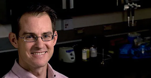 Photo of dark-haired male professor wearing glasses, smiling in lab space with woman pipetting in the background.