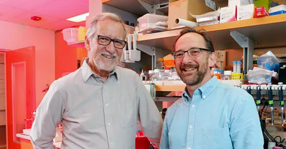 Photo of elder and middle-aged male professors standing in front of shelves with lab equipment and supplies.