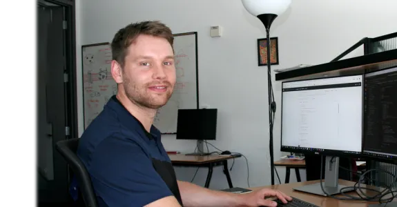 Photo of a white male graduate student wearing a dark blue polo shirt sitting at a beige desk in front of two computer screens. Left screen is white and shows faint lines of code going down the monitor; right screen is black and shows similar lines of code. A whiteboard in the background shows colorful doodles.