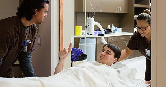 Photo of Andrew Whittaker under sheet on stretcher, giving thumbs up while being wheeled into MRI room by two nurses.