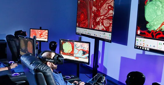 Photo of two surgeons sitting in armchairs, wearing virtual reality headsets in front of large monitors displaying surgery images.