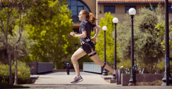 Photo of white female graduate student running across a courtyard area at Stanford, wearing running clothes and the wearable device described in the article.