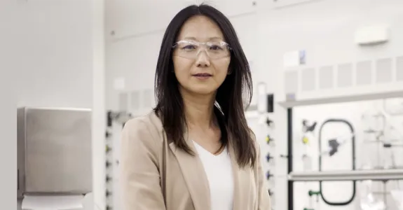 Photo of an Asian female faculty member standing in a very white-toned wet laboratory area, wearing clear protective safety goggles and a beige blazer.