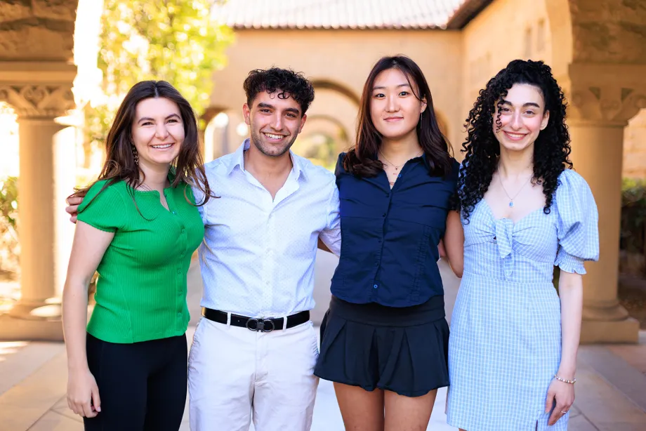 Outdoor photo of 4 undergraduate students standing together, smiling at the camera with their arms around each other.