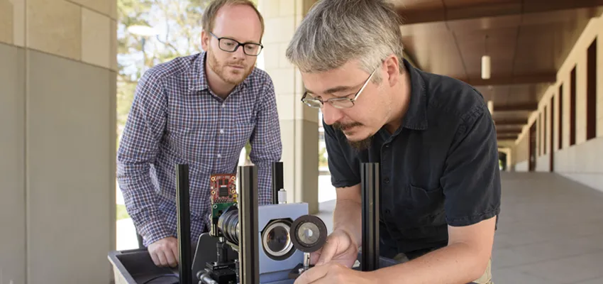 Two men adjusting a camera body resting on a cart; one man is setting up a detached lens near the camera body.
