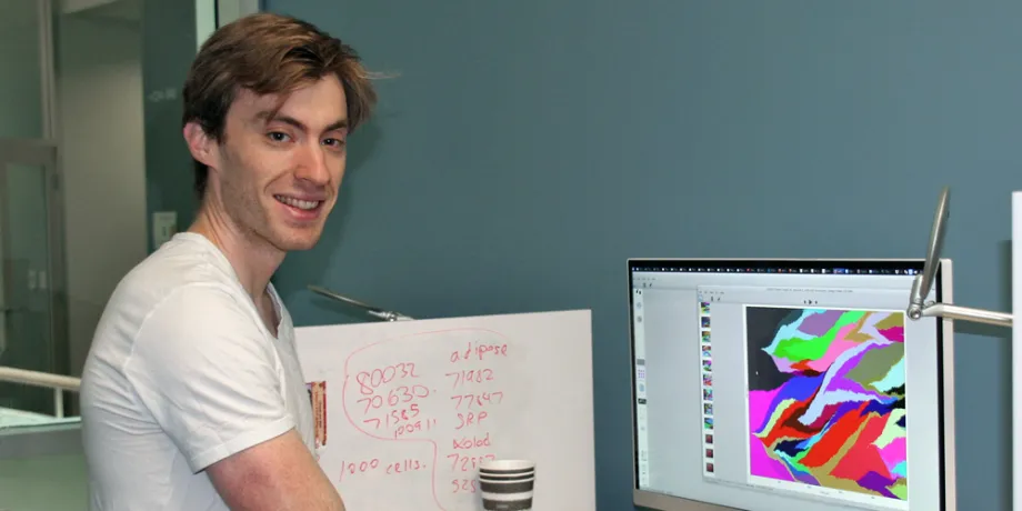 Photo of white male graduate student standing next to a computer displaying colorful data on a large screen.