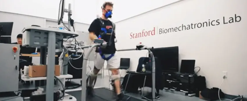 Video screenshot showing a male volunteer wearing ankle exoskeleton devices on a large mechanical treadmill, with the words "Stanford Biomechatronics Lab" on the wall behind.