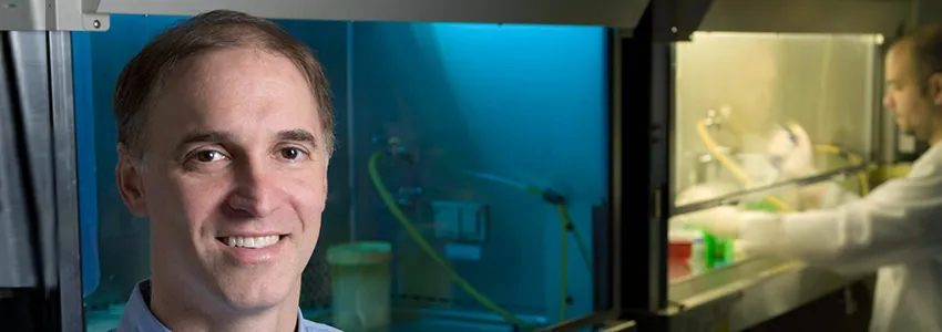 Photo of Dr. Steven Artandi in the lab, with a student working in a fume hood in the background.