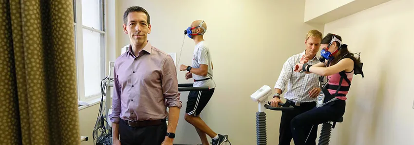 Photo of Dr. Euan Ashley standing at the front of a small room where two subjects, one male and one female, are exercising while wearing multiple kinds of sensors, monitored by a member of the Ashley lab.