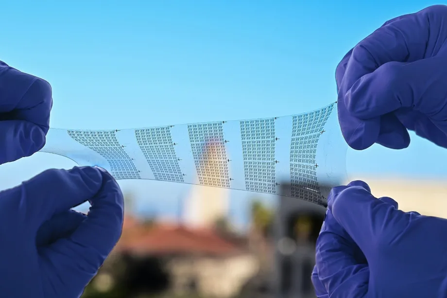 Photo of a transparent film material being stretched and twisted slightly between two pairs of hands wearing gloves. There is a black pattern of electrodes on the clear film.