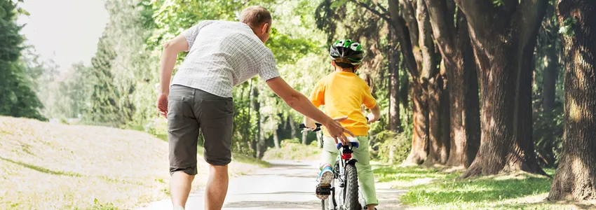 Photo of a father helping his son to learn to ride a bike in a wooded area.