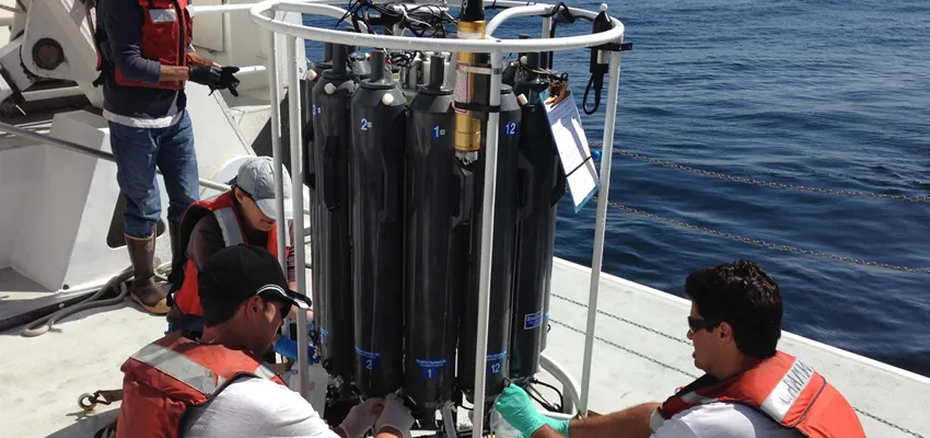 Photo of two men in orange life jackets attending to a sample collector, which is large and cylindrical in shape and comprised of multiple black collection tanks.
