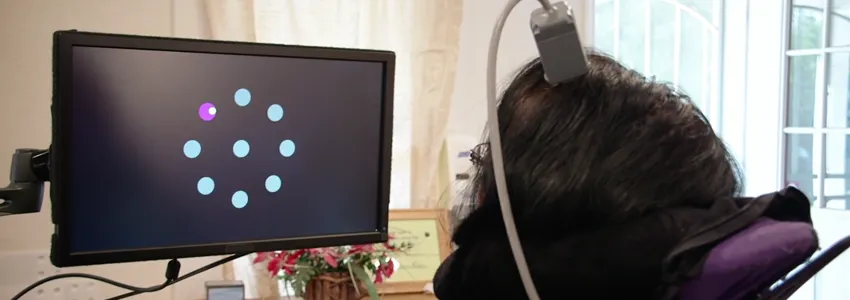 Screenshot from video showing paralyzed woman with device attached to head, using neural signals to manipulate cursor on a screen to select dots lighting up.