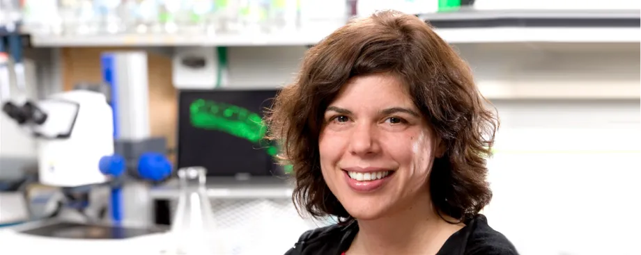 Photo of smiling white female faculty member with shoulder-length wavy brown hair, with a wet lab in the background behind her.