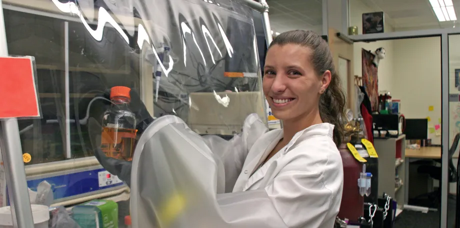 Indoor photo of a white female graduate student with her arms put into a safety hood with plastic, holding up a bottle inside the hood, and smiling at the camera.