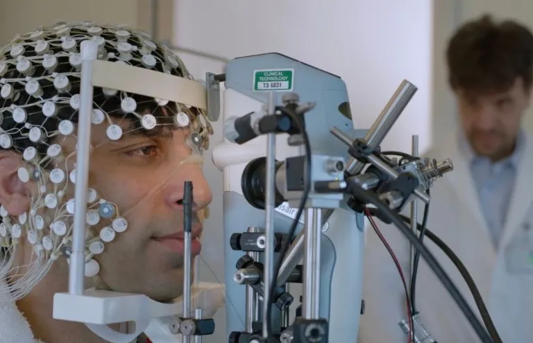 Photo of a man with a net of electrodes on his head with his face resting on a chin rest in front of a machine, with a professor in a lab coat watching from the background.