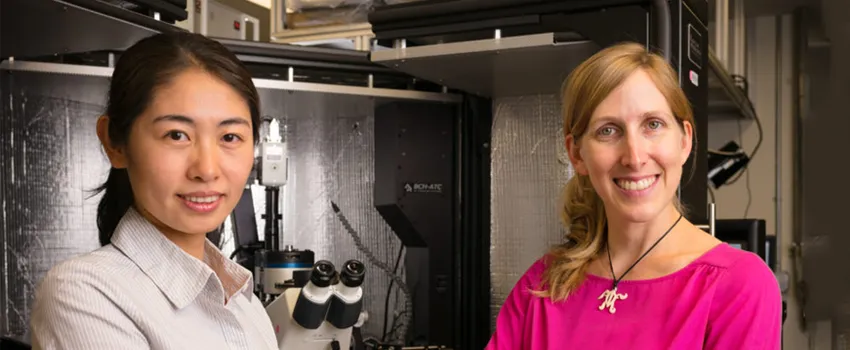 Photo of a young Asian woman and her female Principal Investigator, standing in a specialized lab space with a microscope.