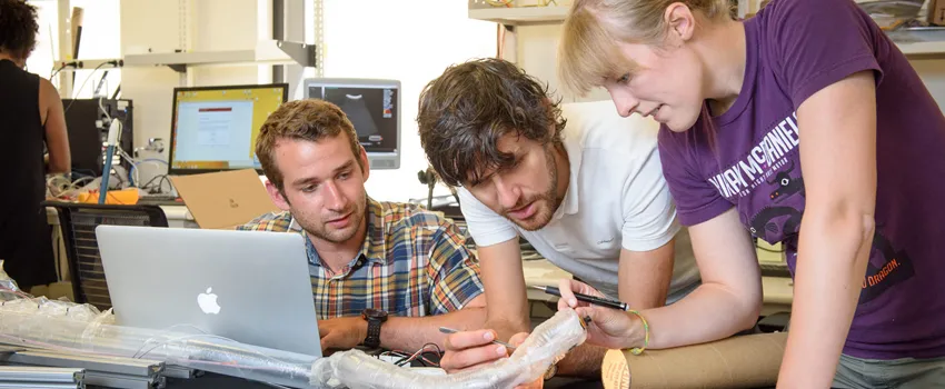 Male and female graduate student work with male visiting professor, consulting a computer screen and examining the translucent "vinebot" robot device.