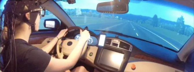 Photo of a young male study participant sitting in a car with a simulated street image past the windshield; participant is wearing a black cap with wires trailing from it and has both hands on the steering wheel.