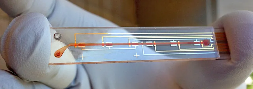 Photo of researcher with gloves on holding up prototype, which looks like a microscope slide with lines on top resembling circuitry, and a channel for blood in the center.