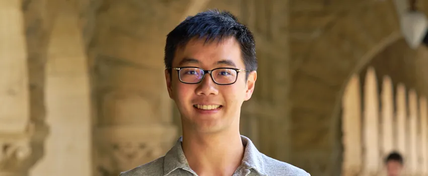 Outdoor headshot photo of a smiling male Asian graduate student with short black hair, wearing glasses.