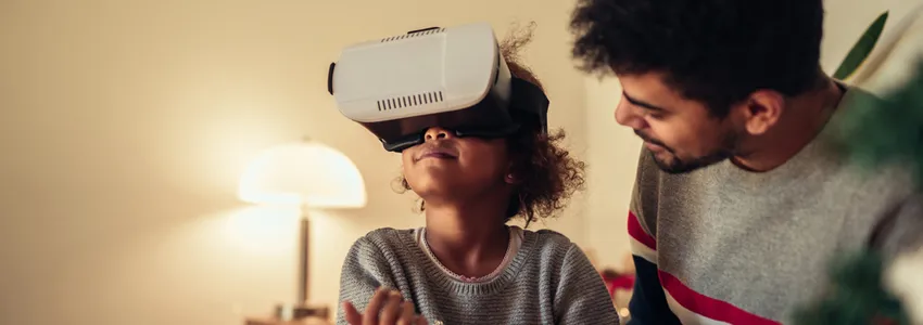 Photo of a young African American girl wearing a VR headset, sitting down with her father.