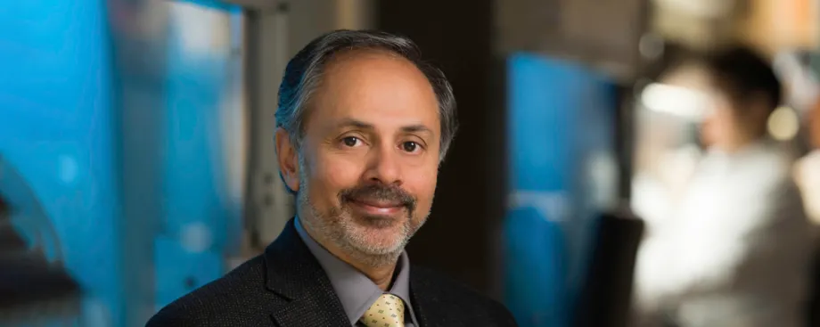 Photo of a smiling man of Indian descent with a beard, wearing a suit and sitting in a laboratory area.