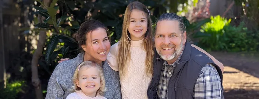 Photo of woman and man with their two young daughters, posed in front of trees.