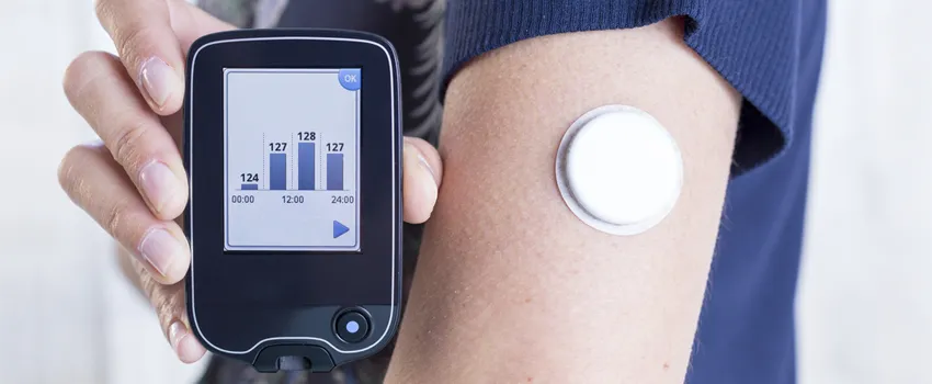 Photo of a woman's arm which bears a glucose monitor, holding up a readout.