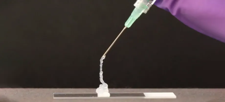 Photograph of a needle extruding a pale translucent gel onto a glass slide, with the photo centered on the needle and showing just a sliver of a purple gloved hand holding it above the slide.