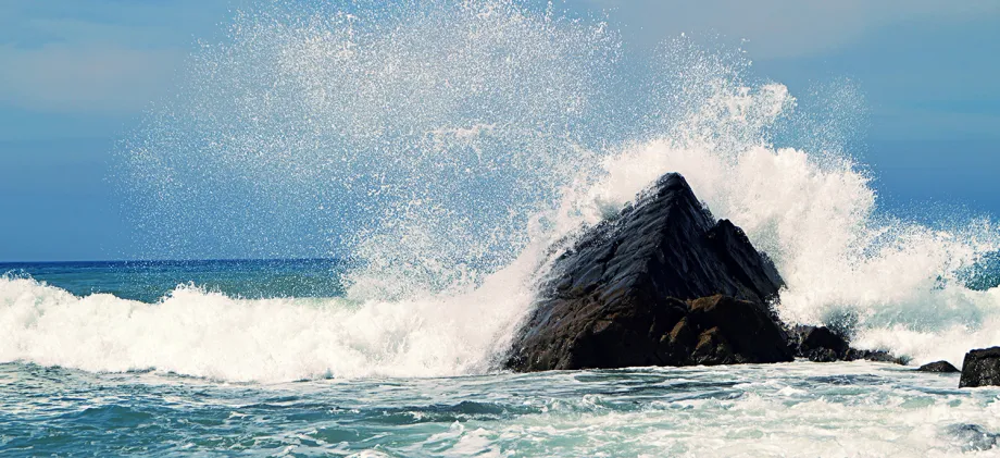 Photo of ocean waves crashing against a large rock and spraying.
