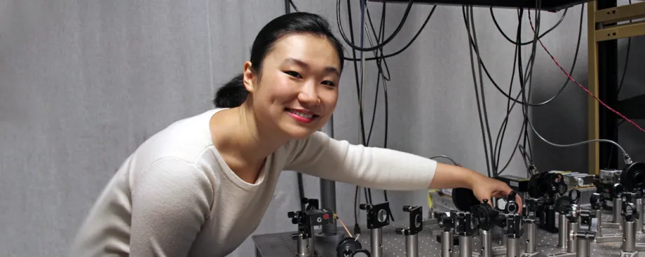 Smiling Asian female graduate student with her hair tied back, wearing a white sweater and leaning down over a complex microscope/laser contraption on a silver table.