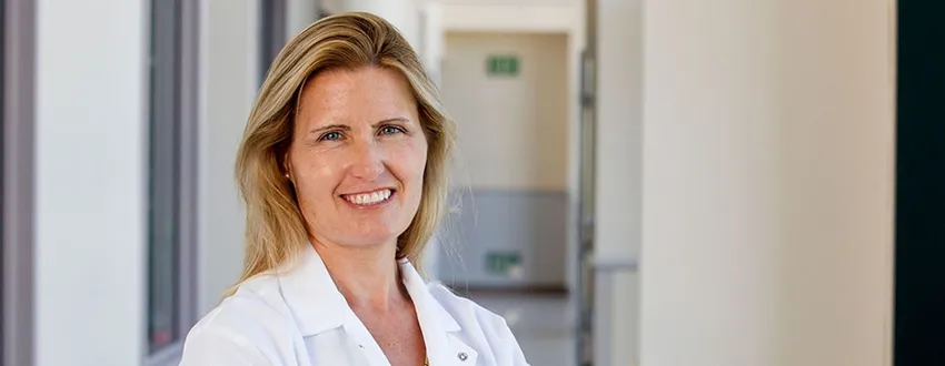 Photo of blonde female PI standing in a hall, smiling, while wearing a white coat.