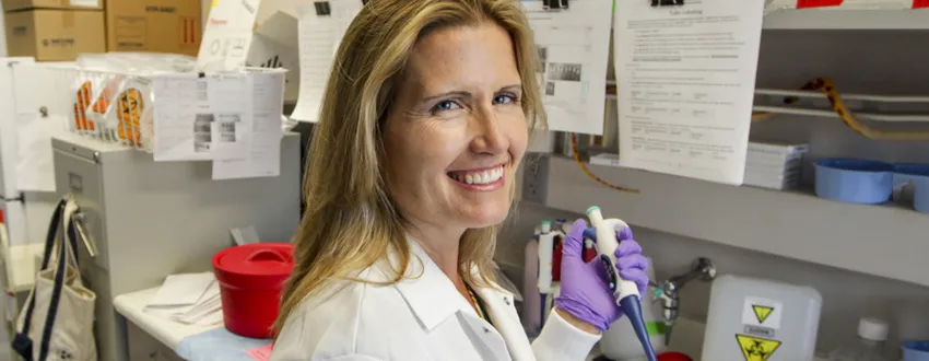 Photo of blonde female PI sitting at bench, using a pipette and smiling.