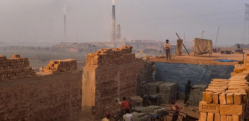 Photo showing workers moving bricks, surrounded by walls made of other bricks, with kilns smoking in the background.