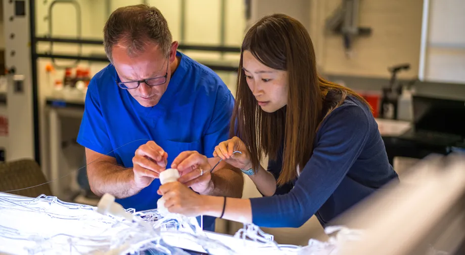 Photo of a male and female scientist both leaned over a plastic device on an illuminated table, adding wires to it.