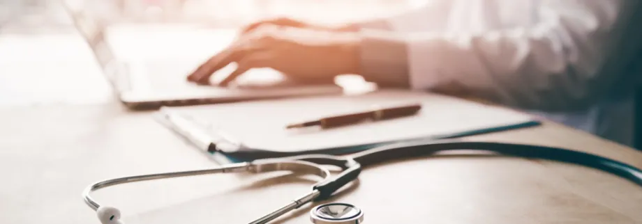 Photo of a doctor's hands using a laptop in the background, with a stethoscope on the table in the foreground.
