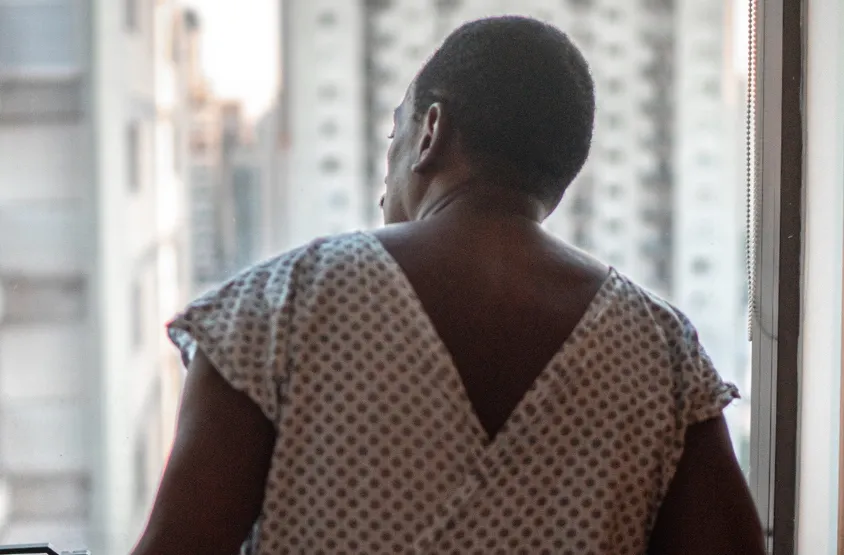 Photo of the back of a man in a hospital gown, in front of a window through which tall buildings are visible.