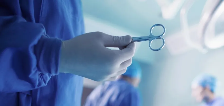 Photo of a surgeon's gloved hand holding up a pair of surgical scissors in the foreground, with operating room activity in the background.