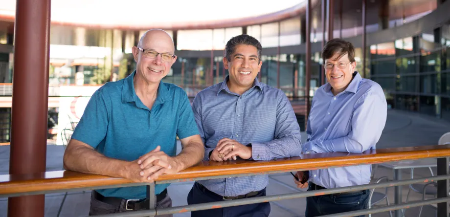 Outdoor photo of Scott Delp, Gabriel Sanchez, and Mark Schnitzer  standing at a railing on an upper level of the Clark Center at Stanford.
