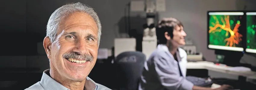 Photo of a middle-aged male PI with a mustache, with a student working on neuroscience displays in the background.