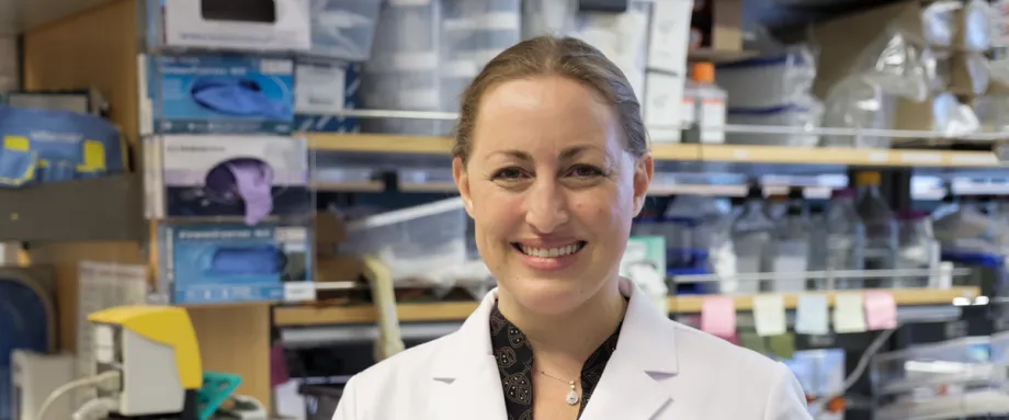 Photo of smiling female faculty member, Dr. Michelle Monje, standing in a wet laboratory space wearing a lab coat. Her blond hair is tied back.