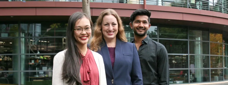Outdoor photo of a young Asian female medical student, a white female faculty member, and a young southeast Asian male undergraduate student smiling at the camera together.