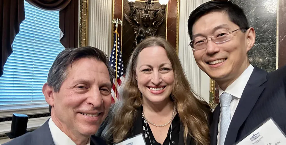 Indoor "selfie" photo of white male faculty member with short gray hair, white female faculty member with long blonde hair, and Asian male faculty member with short black hair and glasses, all smiling at the camera. They are dressed formally, and there are ornate wall decorations showing behind them.
